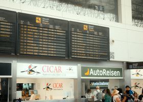 Panel de salidas del aeropuerto sobre los mostradores de Cicar y Autoreisen en una terminal concurrida.
