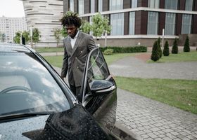Un hombre con un traje gris sale de un coche negro junto a un edificio moderno en una entrada mojada.