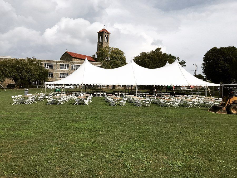 White tents and rows of chairs on a grassy field near a building and clock tower under cloudy skies