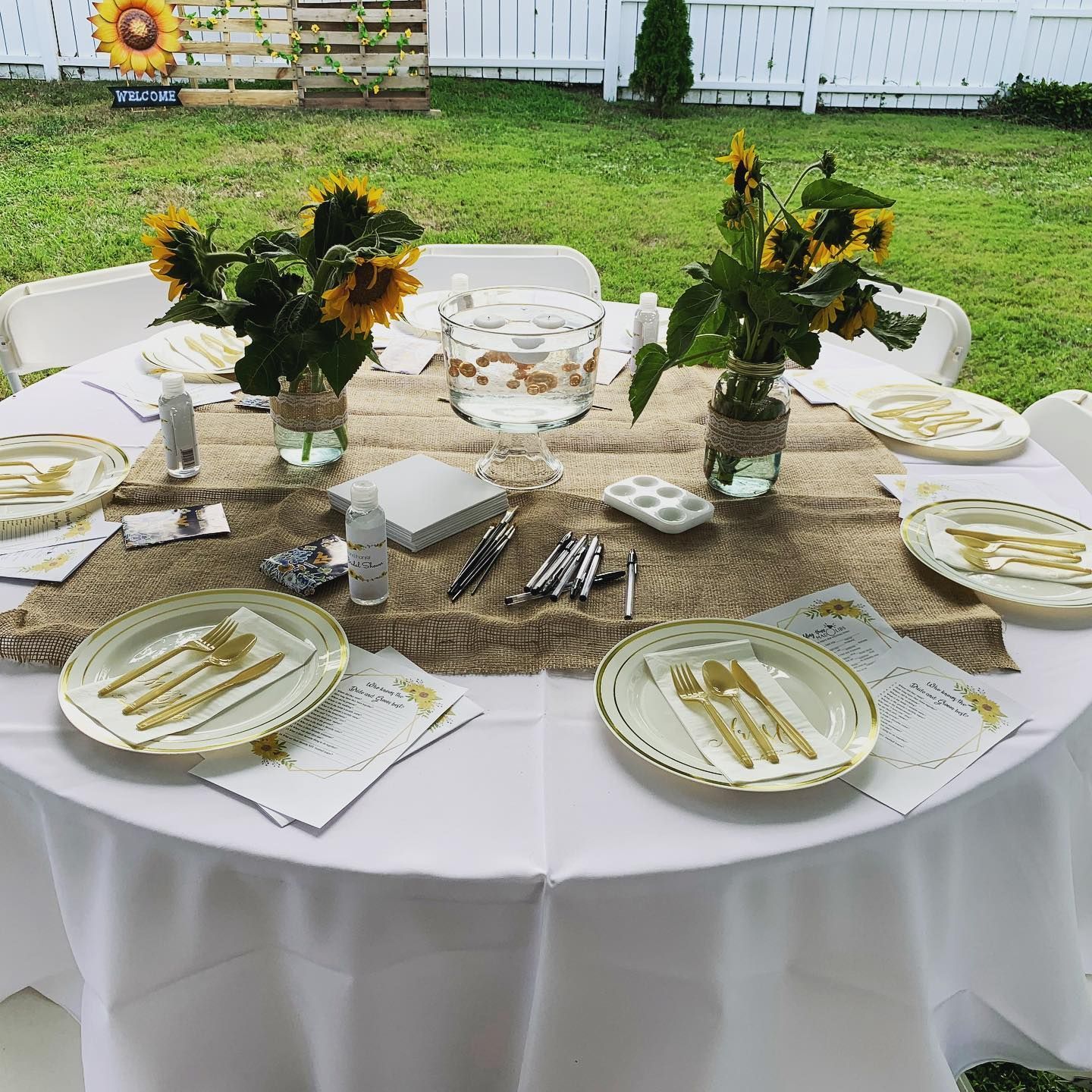 Round outdoor dining table with white tablecloth, gold accents, sunflowers, and place settings on a patio lawn.