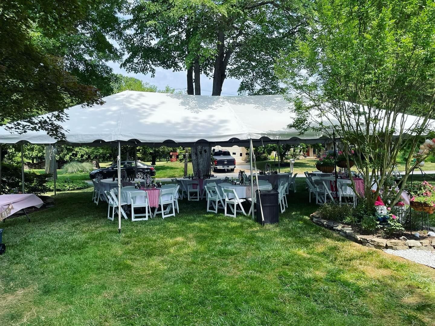 White canopy tent with tables and chairs set up on a grassy lawn under trees, with flowers nearby