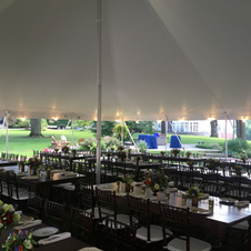 Outdoor tented banquet setup with long tables, black chairs, and a green lawn beyond.