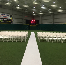 Indoor event hall with rows of white chairs, a white aisle, green walls, and a digital scoreboard at the far end