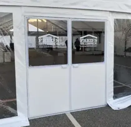 White double-door entrance to a tented building with windows and parked cars reflected in the glass.