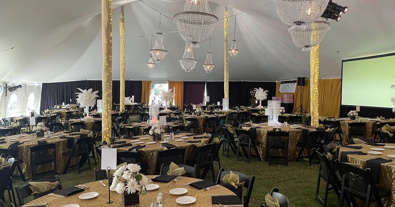 Decorated banquet hall with round tables, gold chairs, chandeliers, and white floral centerpieces under a tented ceiling