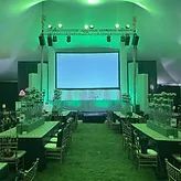 Green-lit conference tent with rows of tables and chairs facing a large projection screen