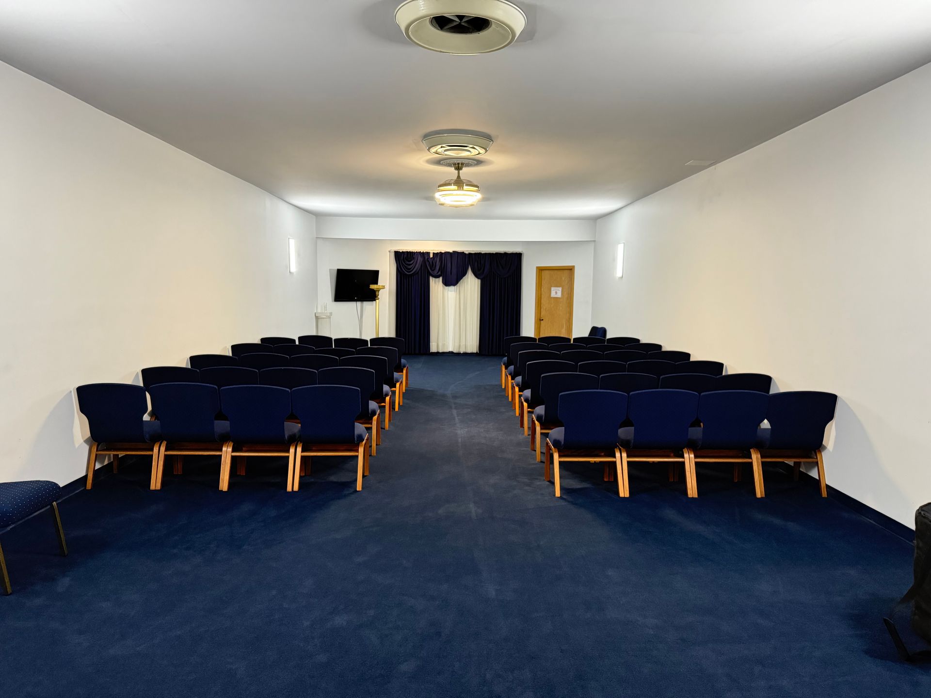 Rows of dark blue chairs face a small stage draped with navy blue curtains in a brightly lit auditorium.