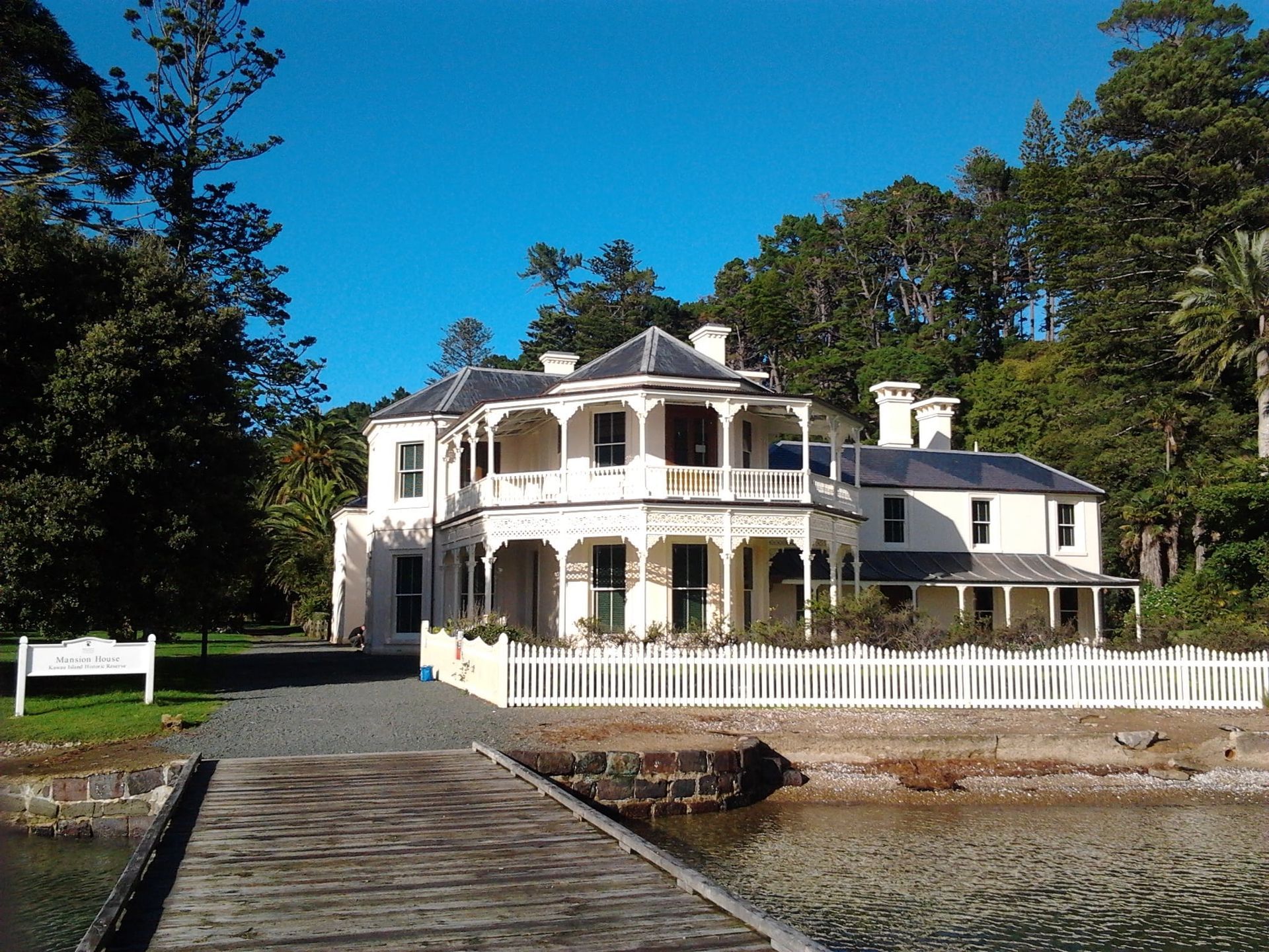 Two-story white house with balcony, white picket fence, next to a pier and water, under a blue sky.