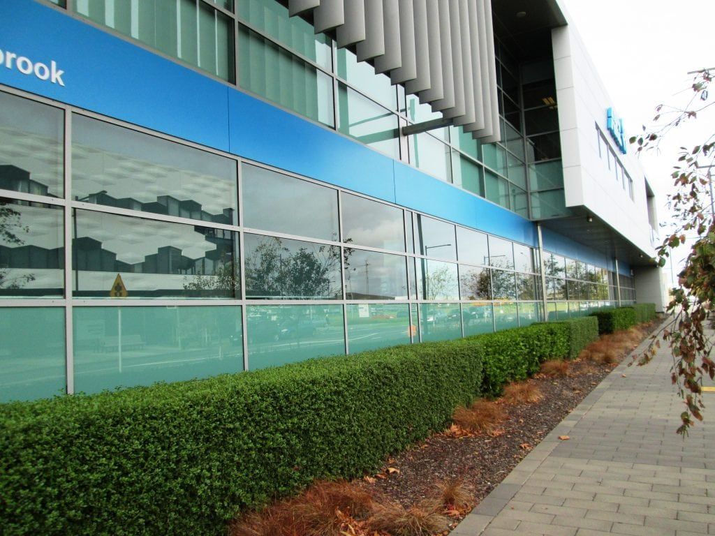Blue and glass building facade with reflective windows, green hedge, and gray sidewalk.