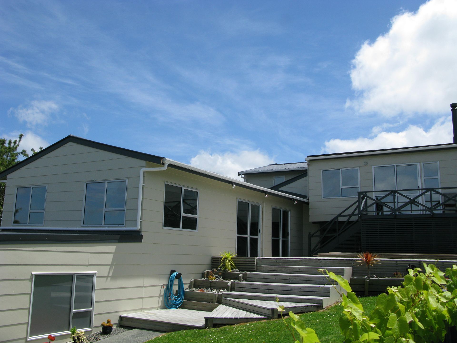 Light-colored house with multiple sections and decks against a blue sky. Green grass and shrubs in the foreground.