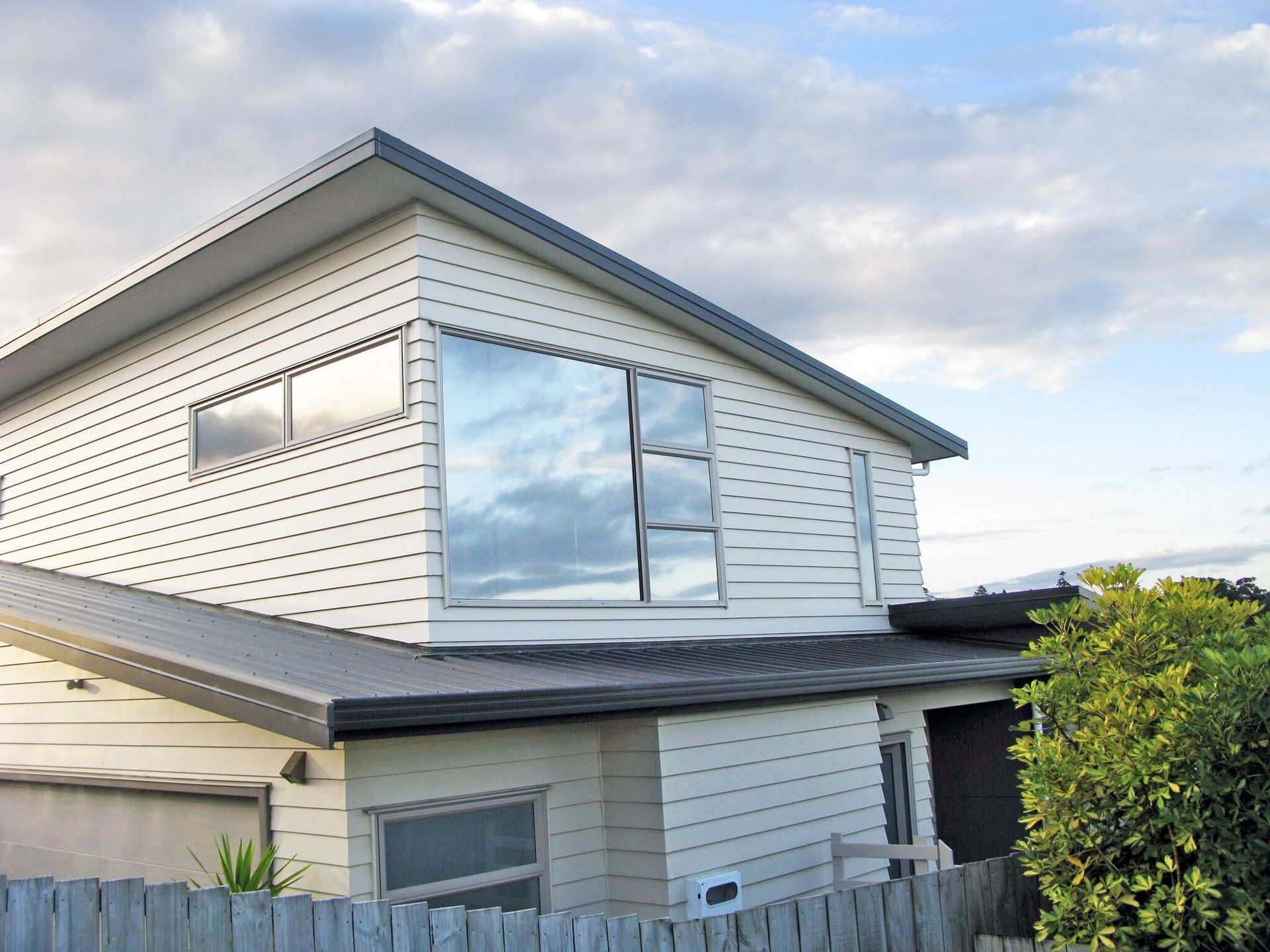 Two-story white house with large reflective windows, gray roof, and a wooden fence. Cloudy sky.