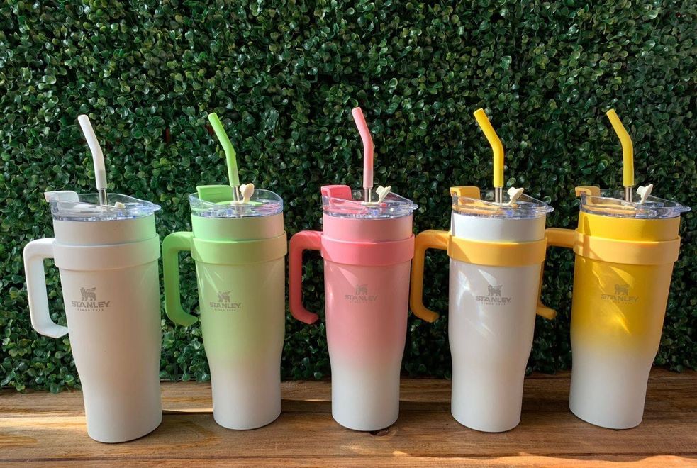 Cinco bebidas heladas en tonos pastel, en tazas con asa y pajitas, alineadas sobre una mesa de madera frente a un seto verde