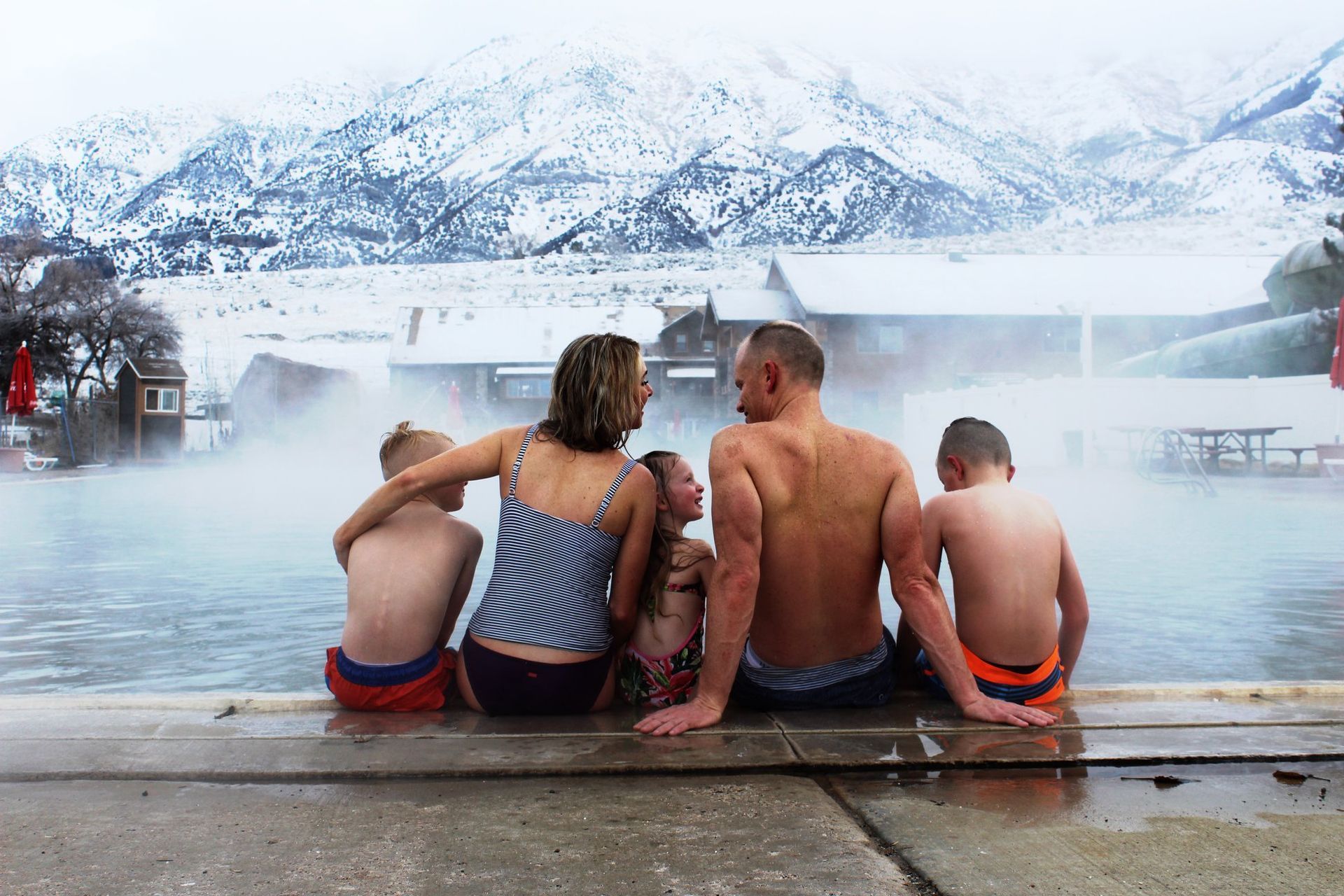 Family in swimwear sitting by a steaming pool with snow-covered mountains in the background.