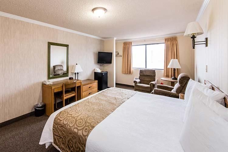A hotel room with a queen bed, a desk, a mirror, a television, and two armchairs near a window with curtains.