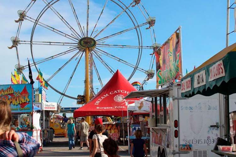 A Ferris wheel rises above a fairground midway with food stalls, tents, and people walking on a sunny day.