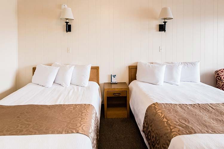 Two hotel beds with white linens and tan runners, separated by a wooden nightstand with wall lamps against a beige wall.