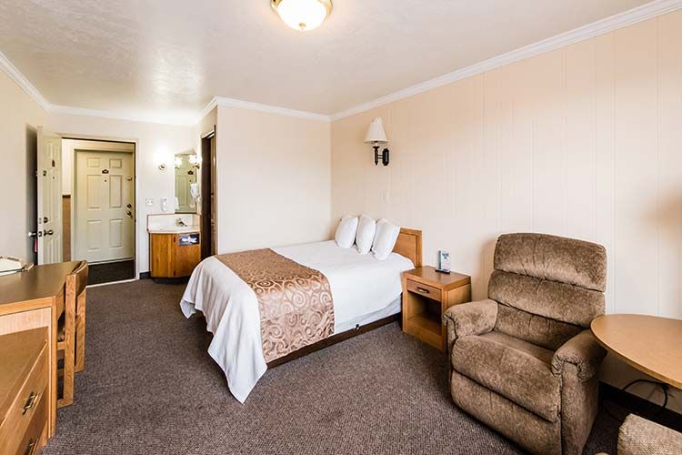 A hotel room with a made bed, a tan recliner, a wooden desk, and an entryway leading to a bathroom.