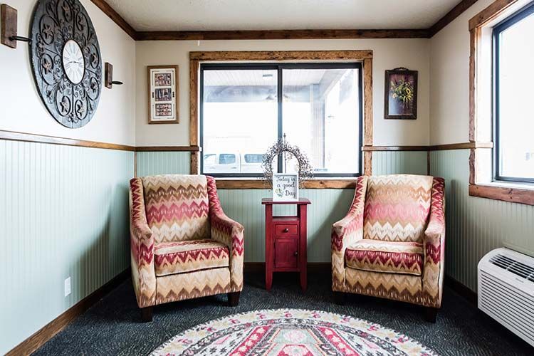 Two patterned armchairs flanking a small red table in front of a window, inside a room with sage green wainscoting.