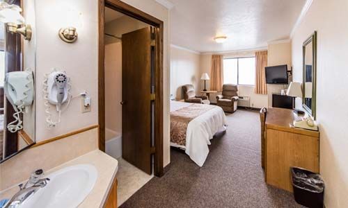 Hotel room interior featuring a vanity area with a hair dryer, a bathroom, a bed, seating, and a desk with a television.