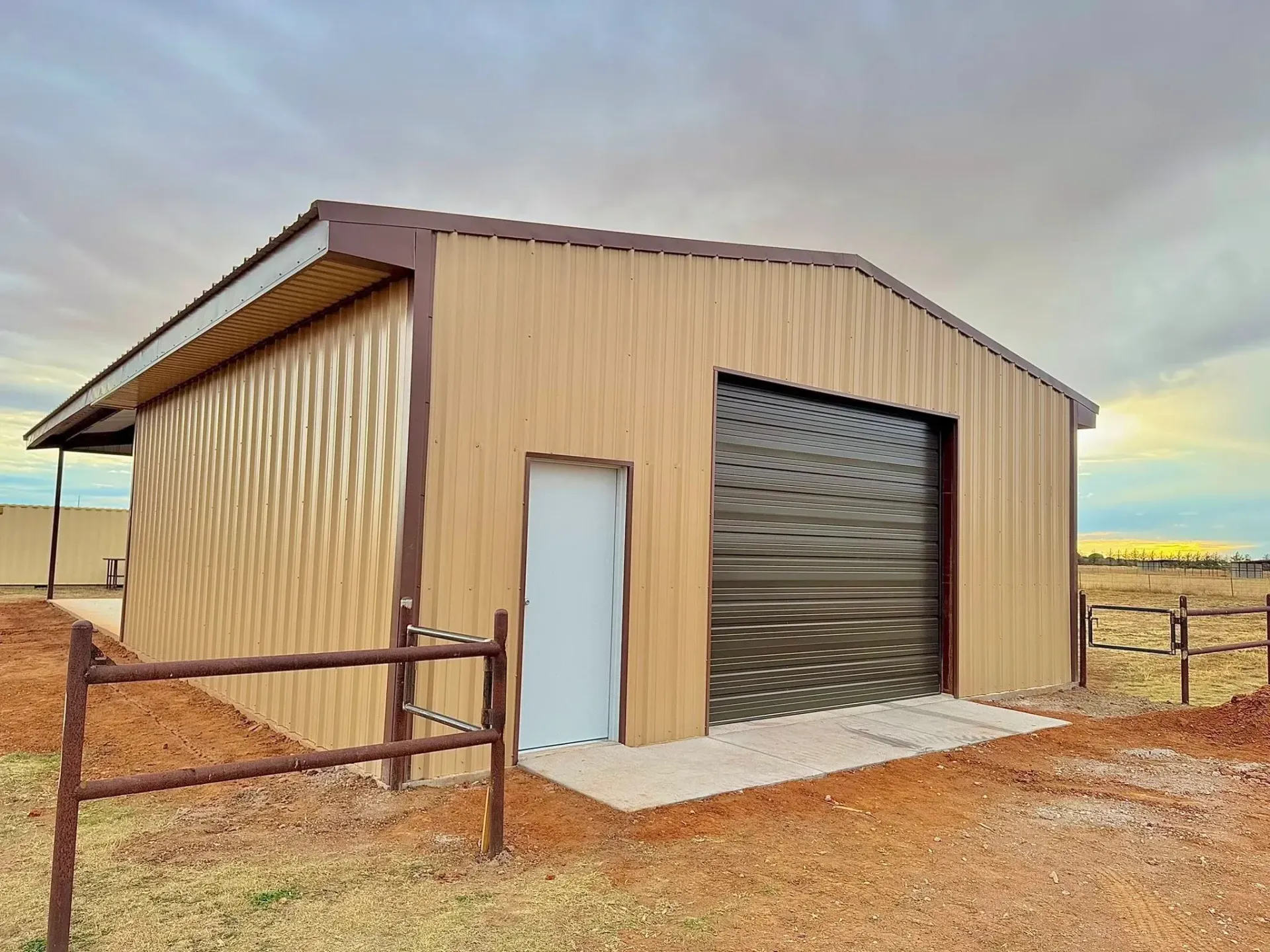 Small tan metal shed with a white door and large roll-up door in a gravel lot at sunset.