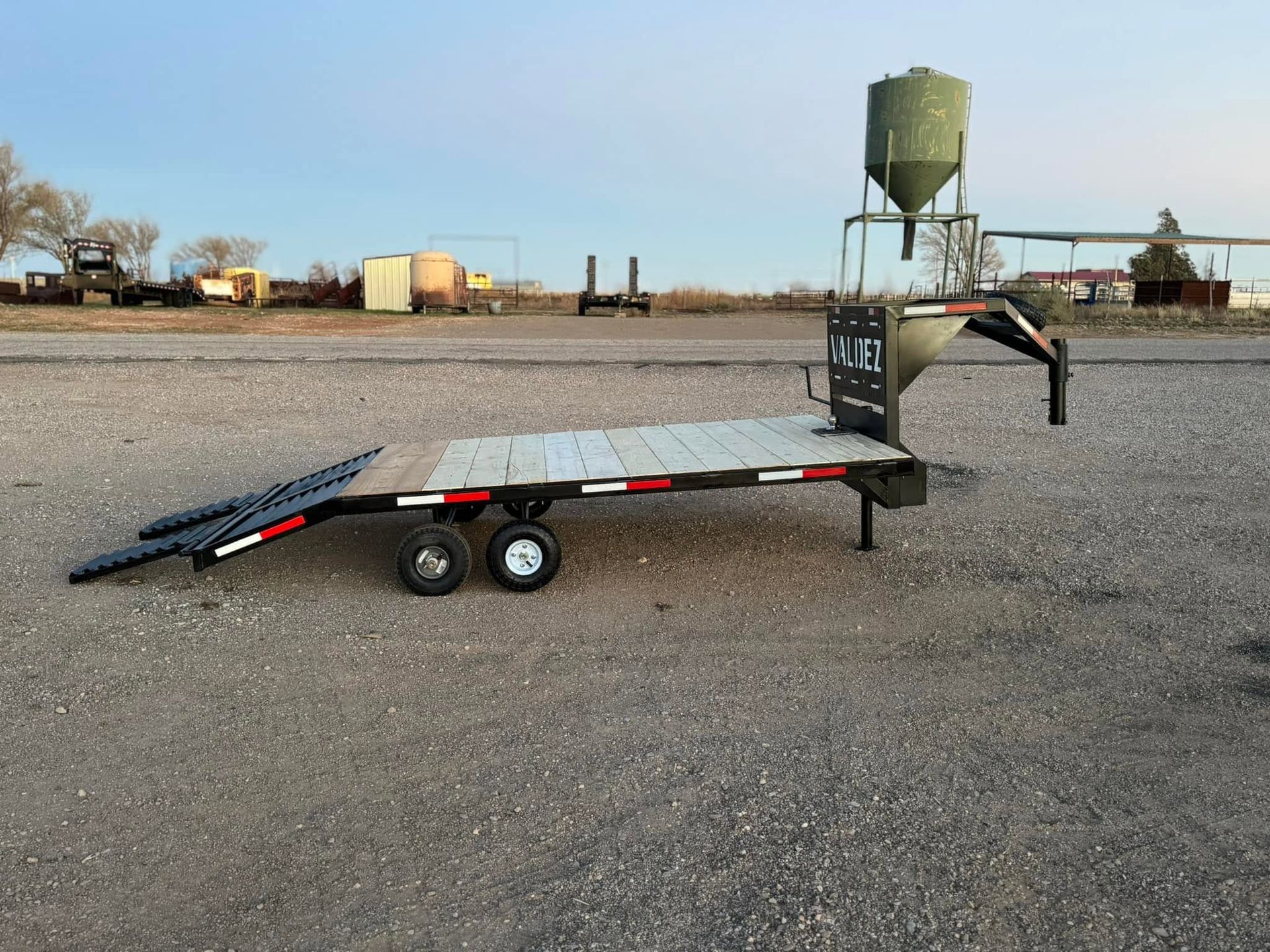 Black flatbed trailer in a gravel lot with a green grain silo in the background.