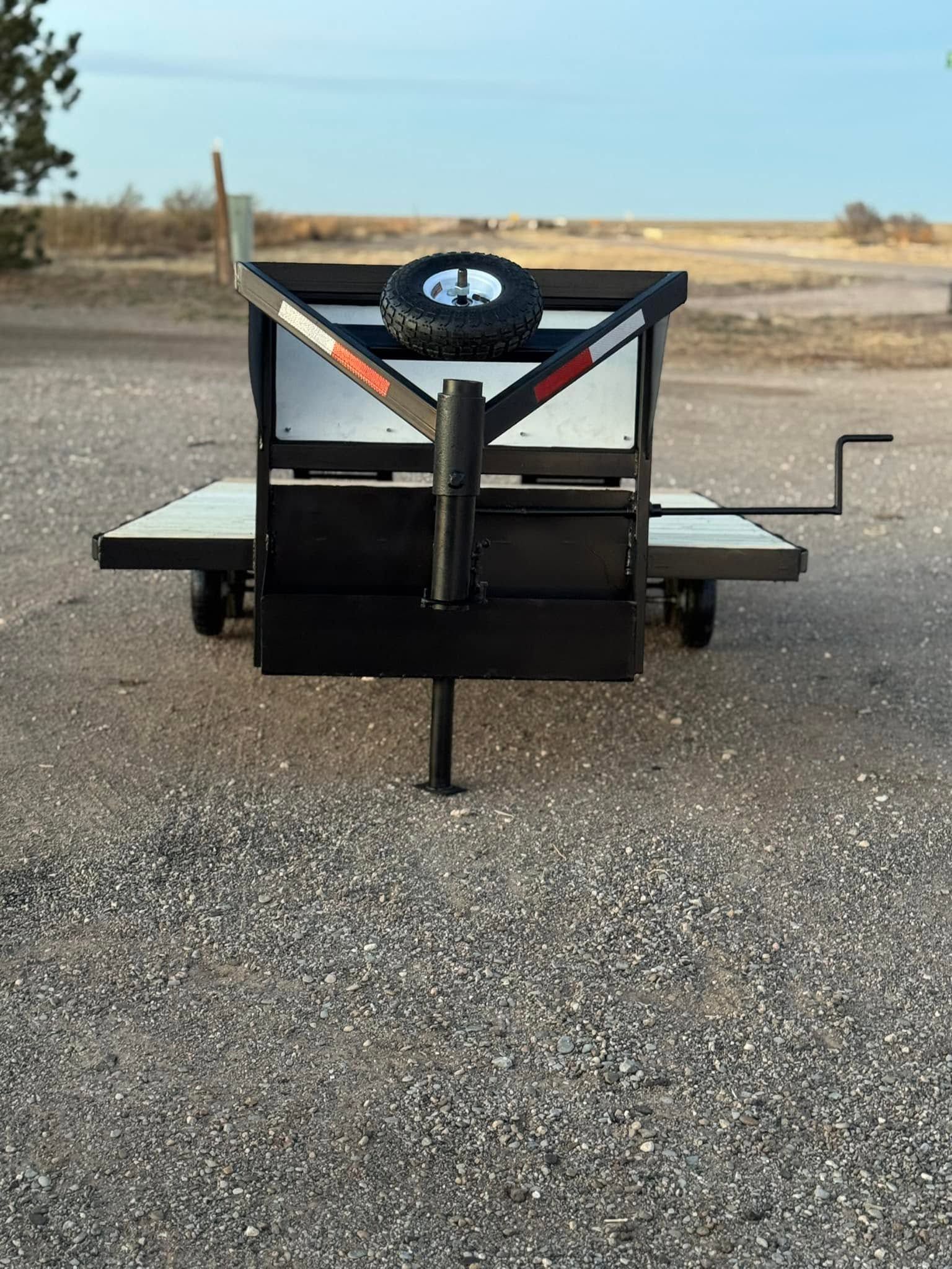 Black utility trailer with side ramps and spare tire, parked on gravel in an open field.