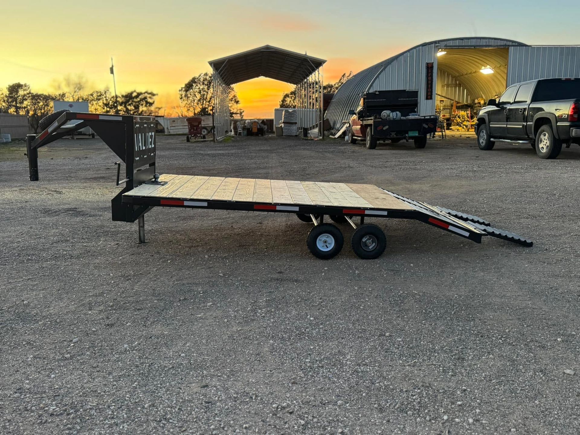 Flatbed trailer with ramp in a gravel lot at sunset, with trucks and metal sheds in the background