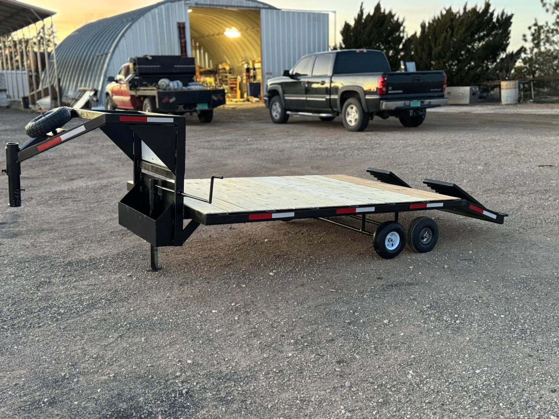 Black equipment trailer with ramp parked on gravel near a metal building and pickup truck