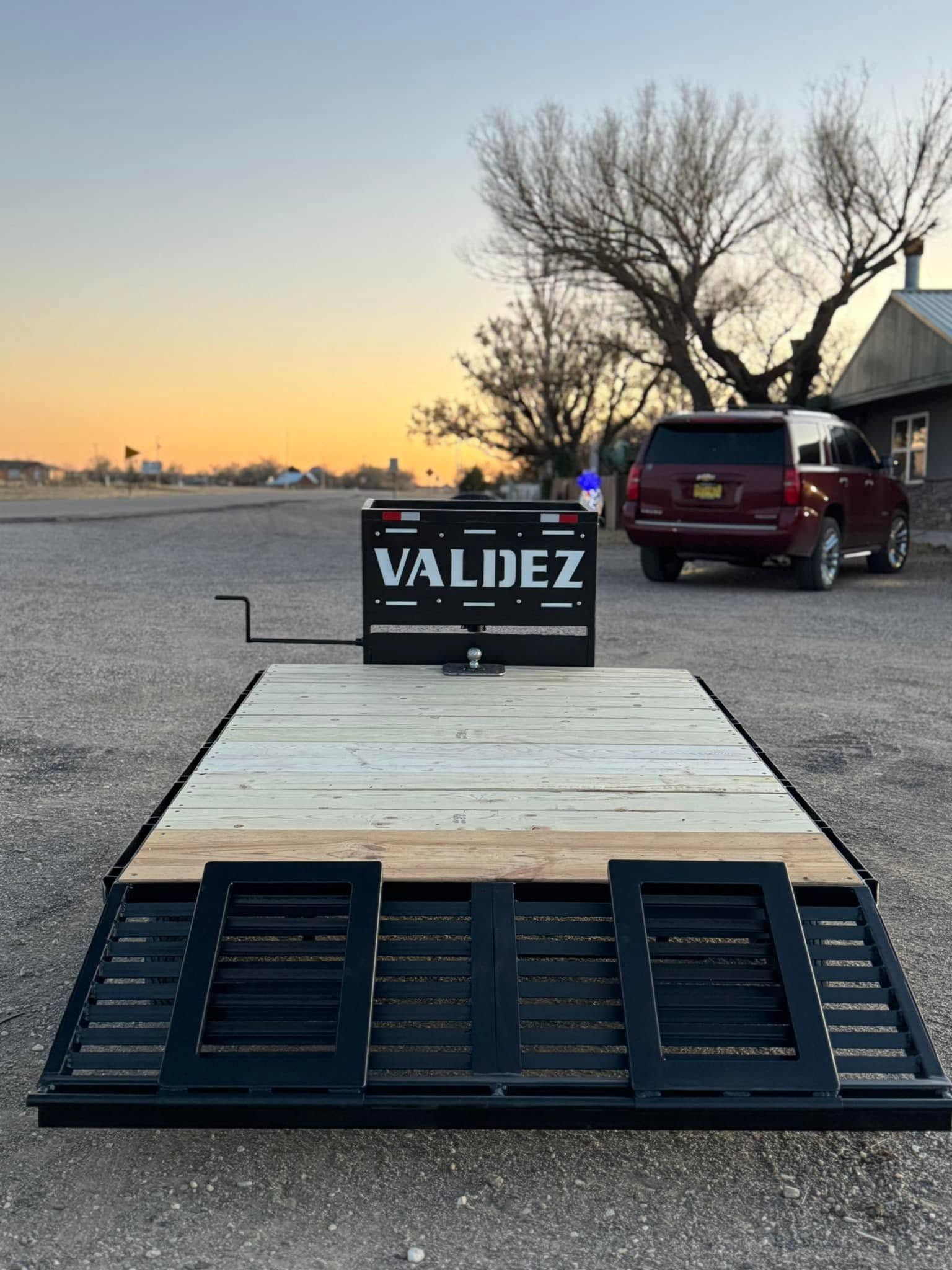 Black utility trailer ramp with a Valiez logo, parked on a roadside at sunset with trucks in the background