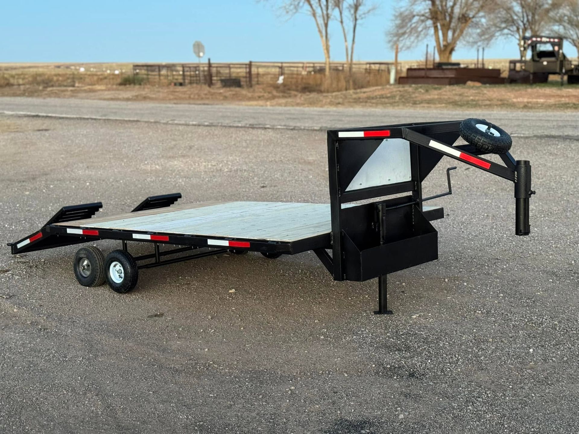 Black utility trailer with ramp and spare tire parked on a gravel lot under a clear sky.