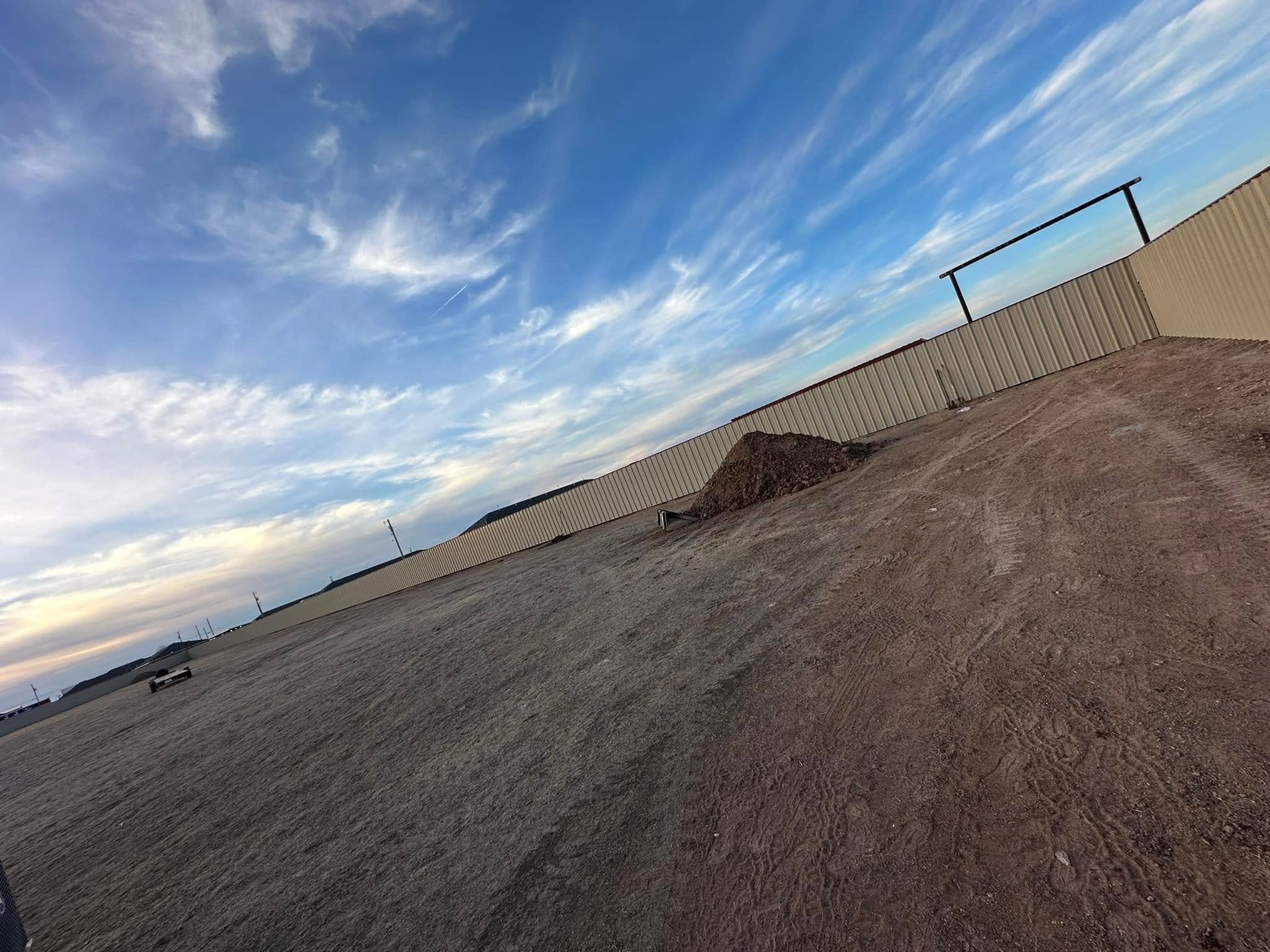 Tilted view of a dirt lot beside a tall fence under a blue, cloud-streaked sky