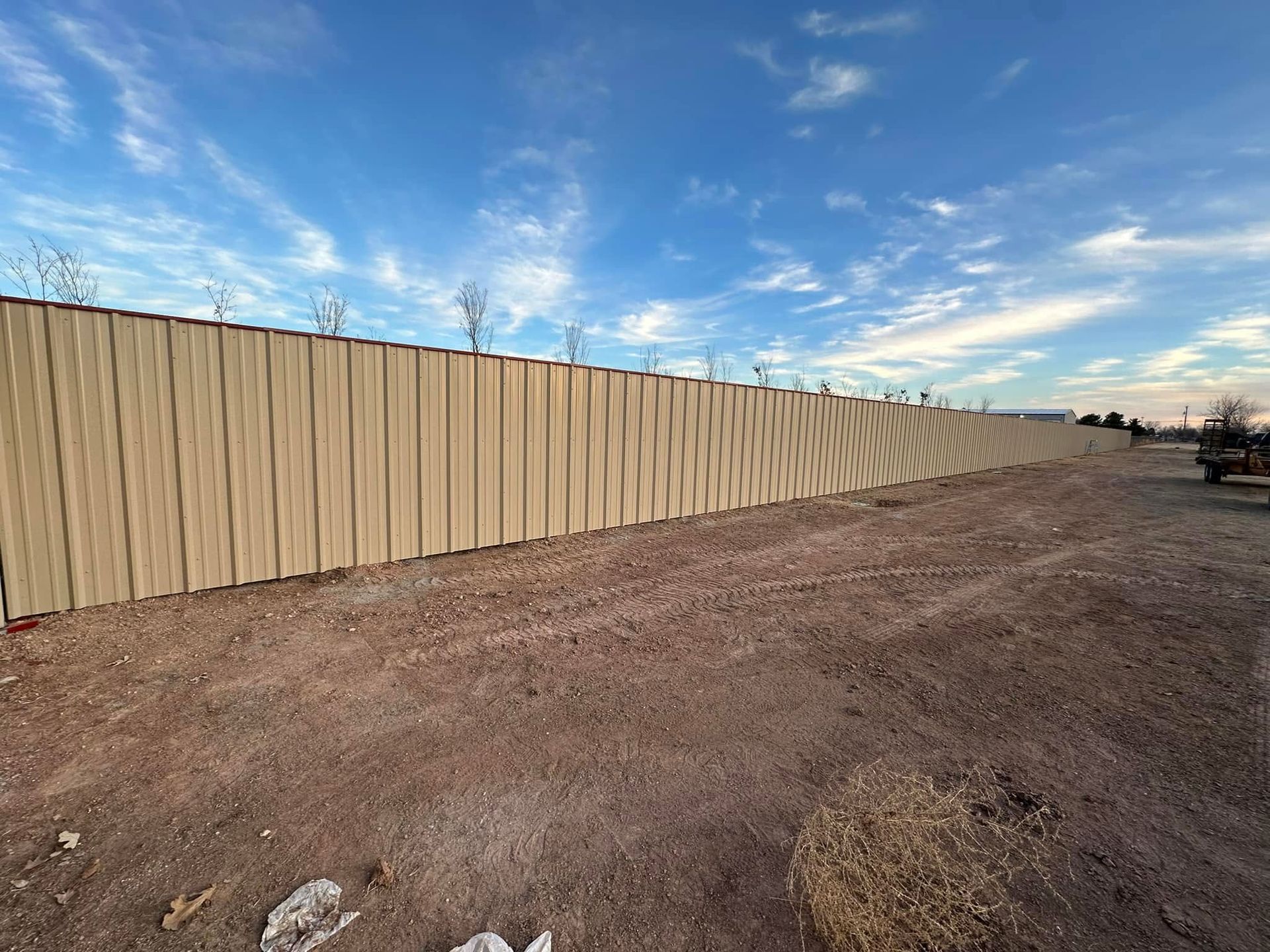 Long tan fence along a barren dirt lot under a blue, cloud-streaked sky