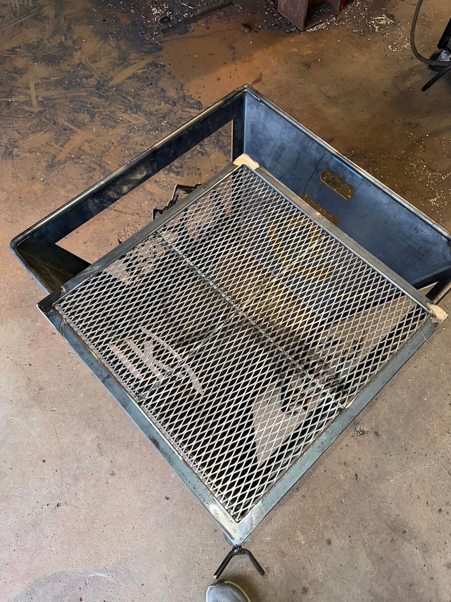 Metal mesh fire pit or grill basket on a concrete floor, with blue sides and a handle.