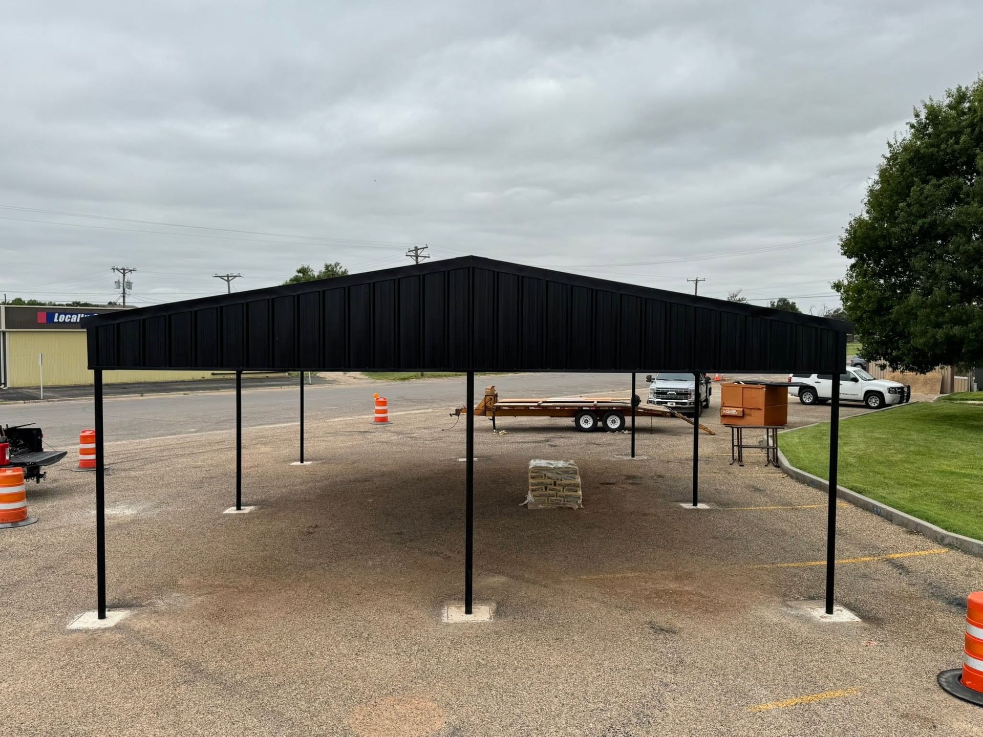 Black canopy tent on poles over a gravel lot, with orange cones and a cloudy sky.