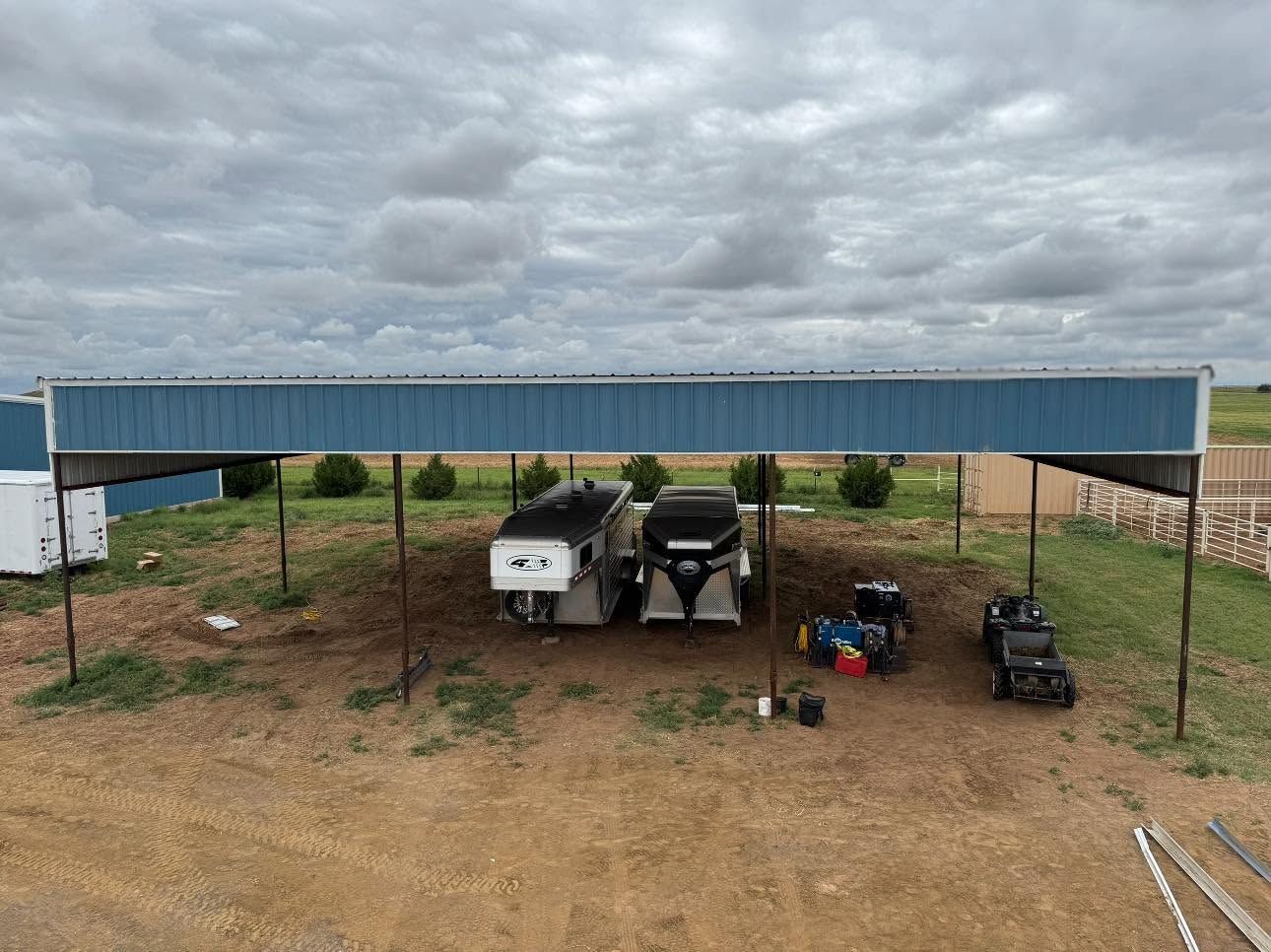 Blue-roofed metal shelter over parked equipment and vehicles on a gravel lot under cloudy skies