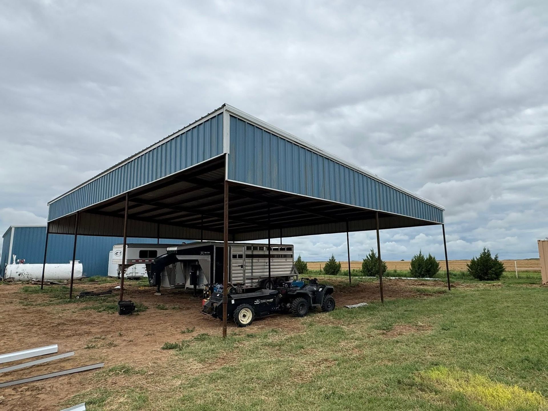 Blue metal open-sided shed with vehicles parked underneath on a cloudy rural property