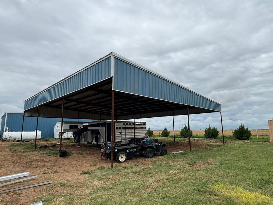Metal carport shelter on a rural lot, with vehicles and equipment underneath under a cloudy sky.