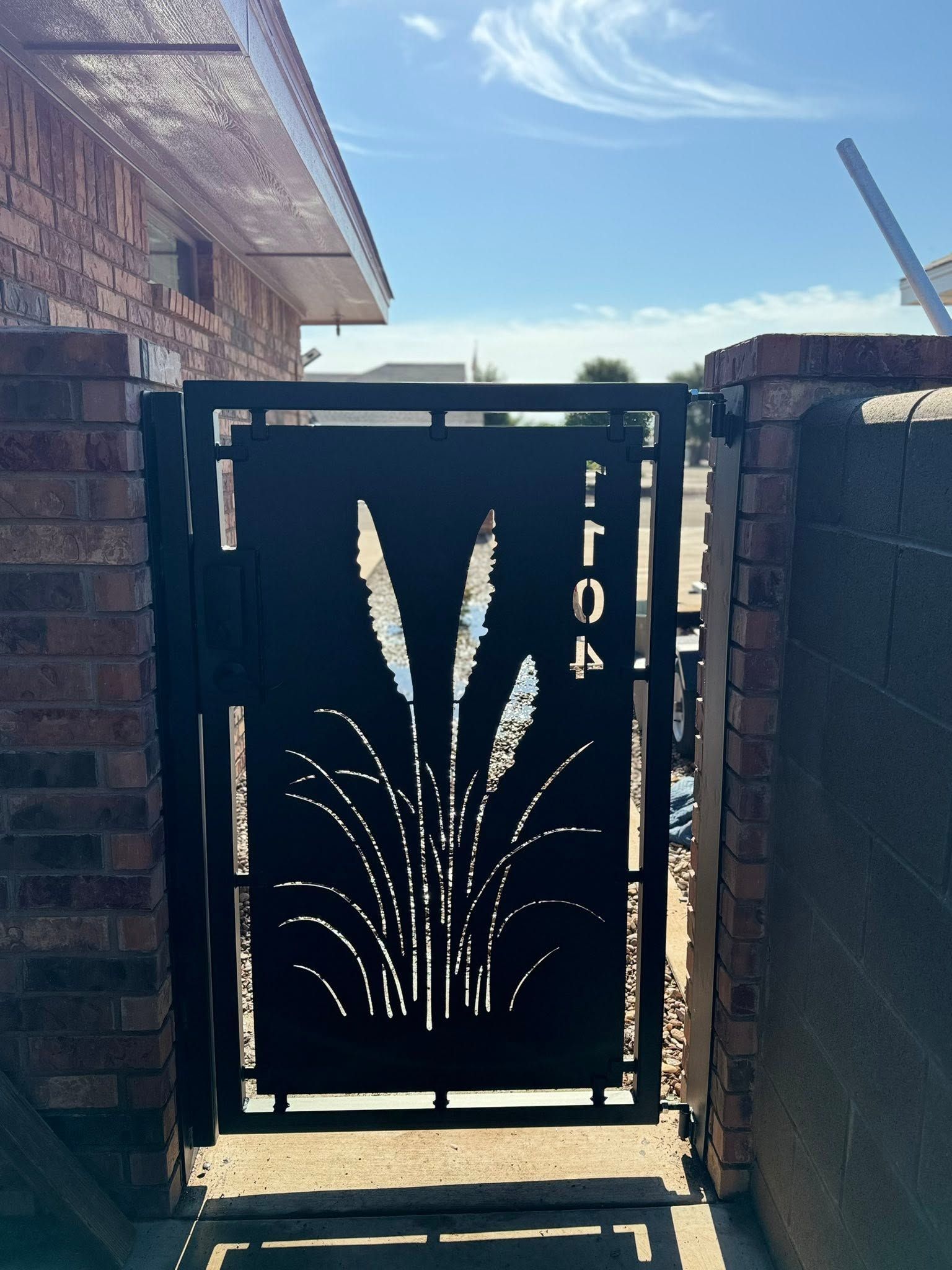 Black metal gate with decorative grass design between brick walls, viewed in bright daylight.