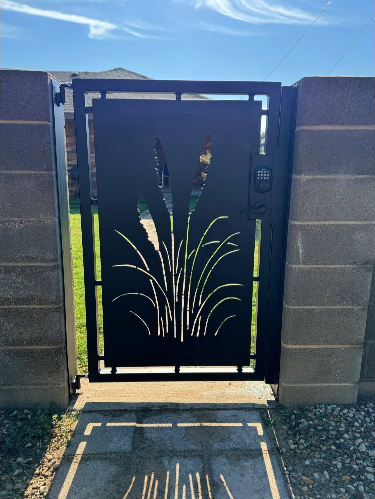 Black metal gate with decorative grass cutouts between cinder block walls under a blue sky