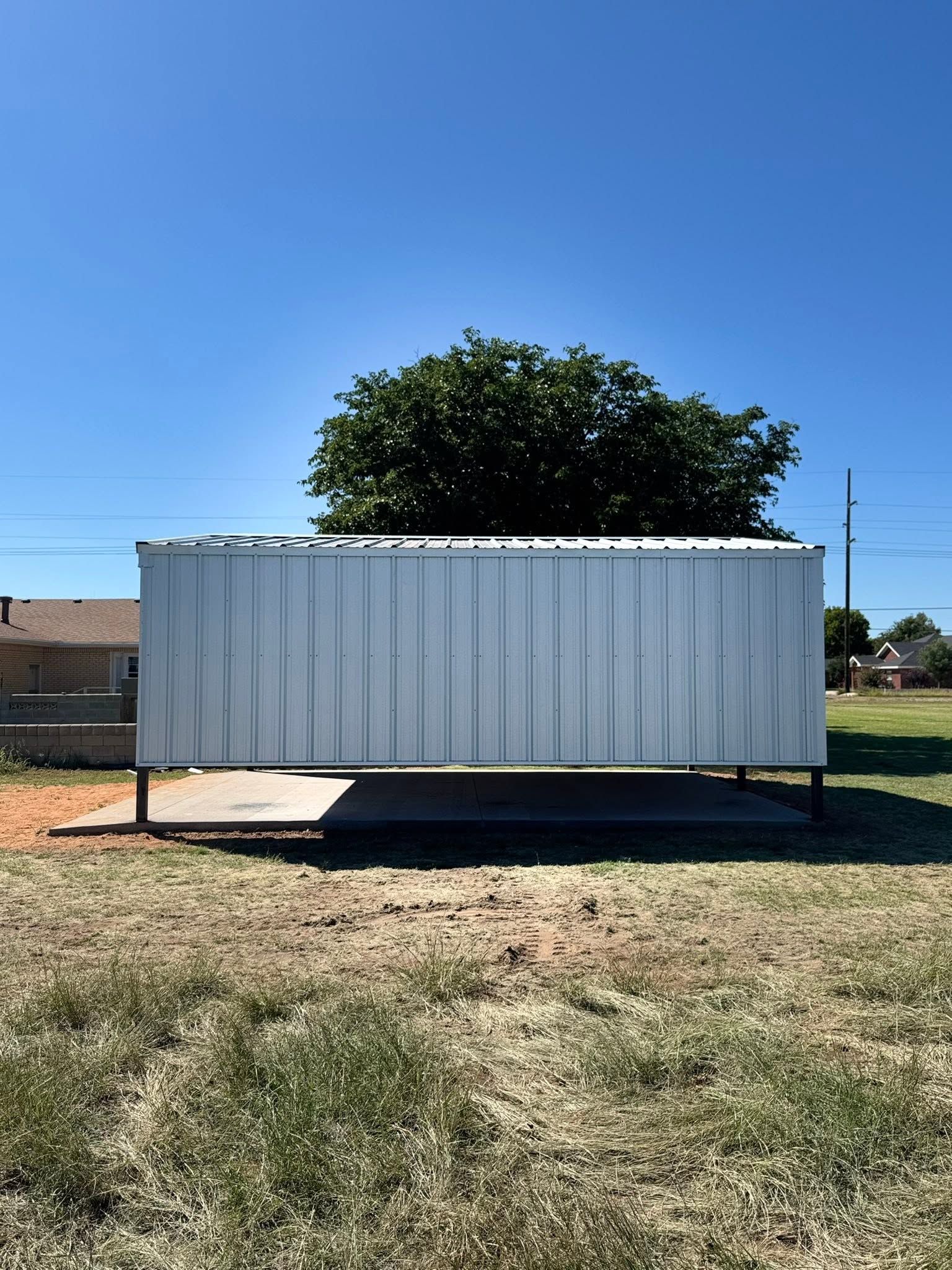 White corrugated metal shed on a grassy lot under a clear blue sky, with a tree behind it.