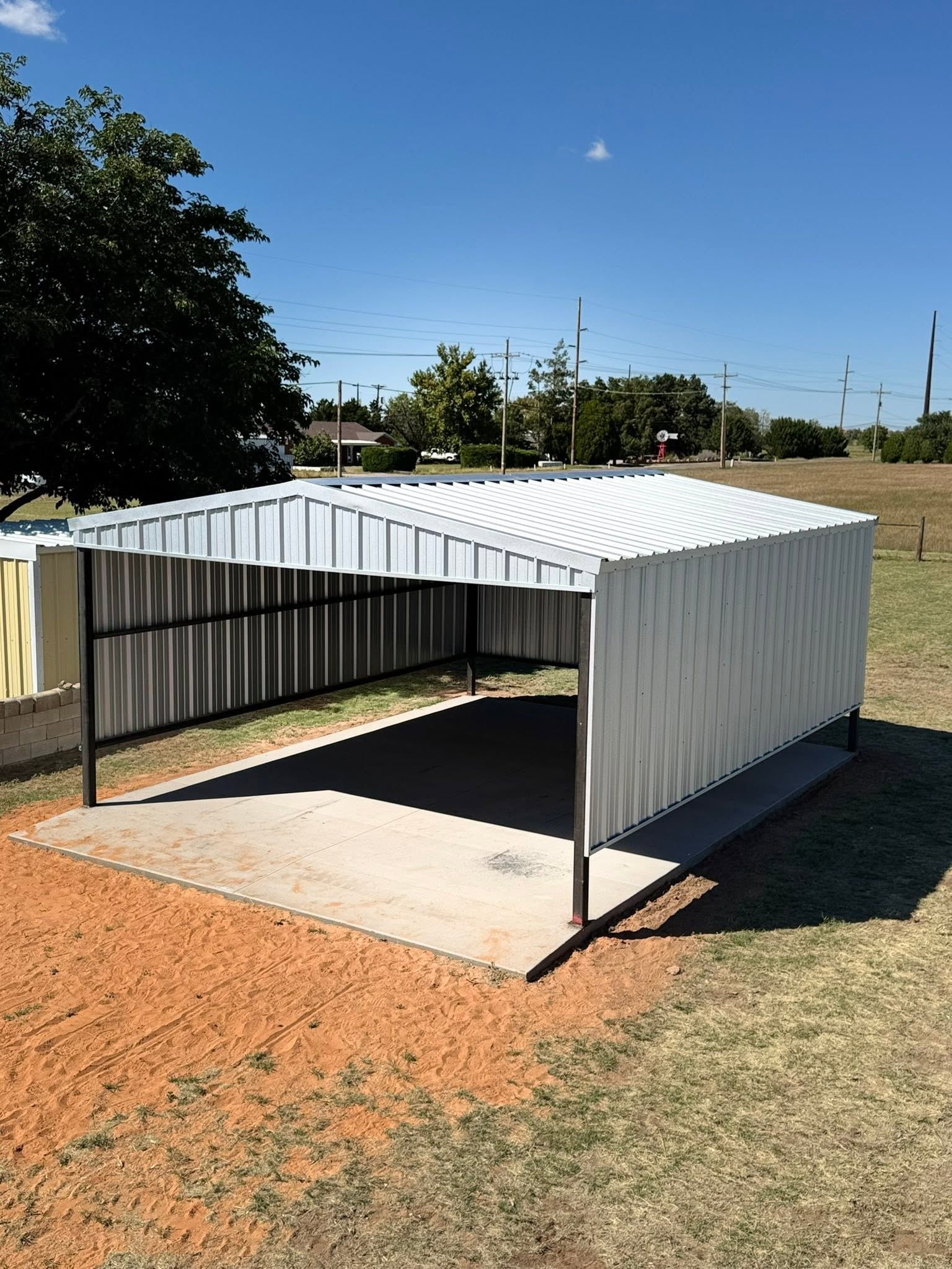 White corrugated metal carport on a concrete pad beside a fenced yard under a clear blue sky