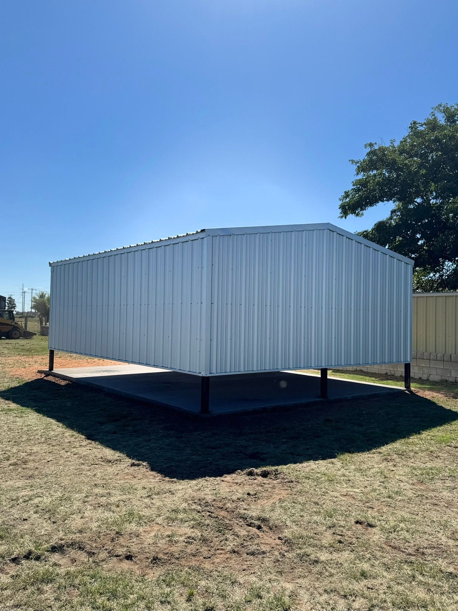 White corrugated metal shed raised on stilts in a grassy field under a blue sky.