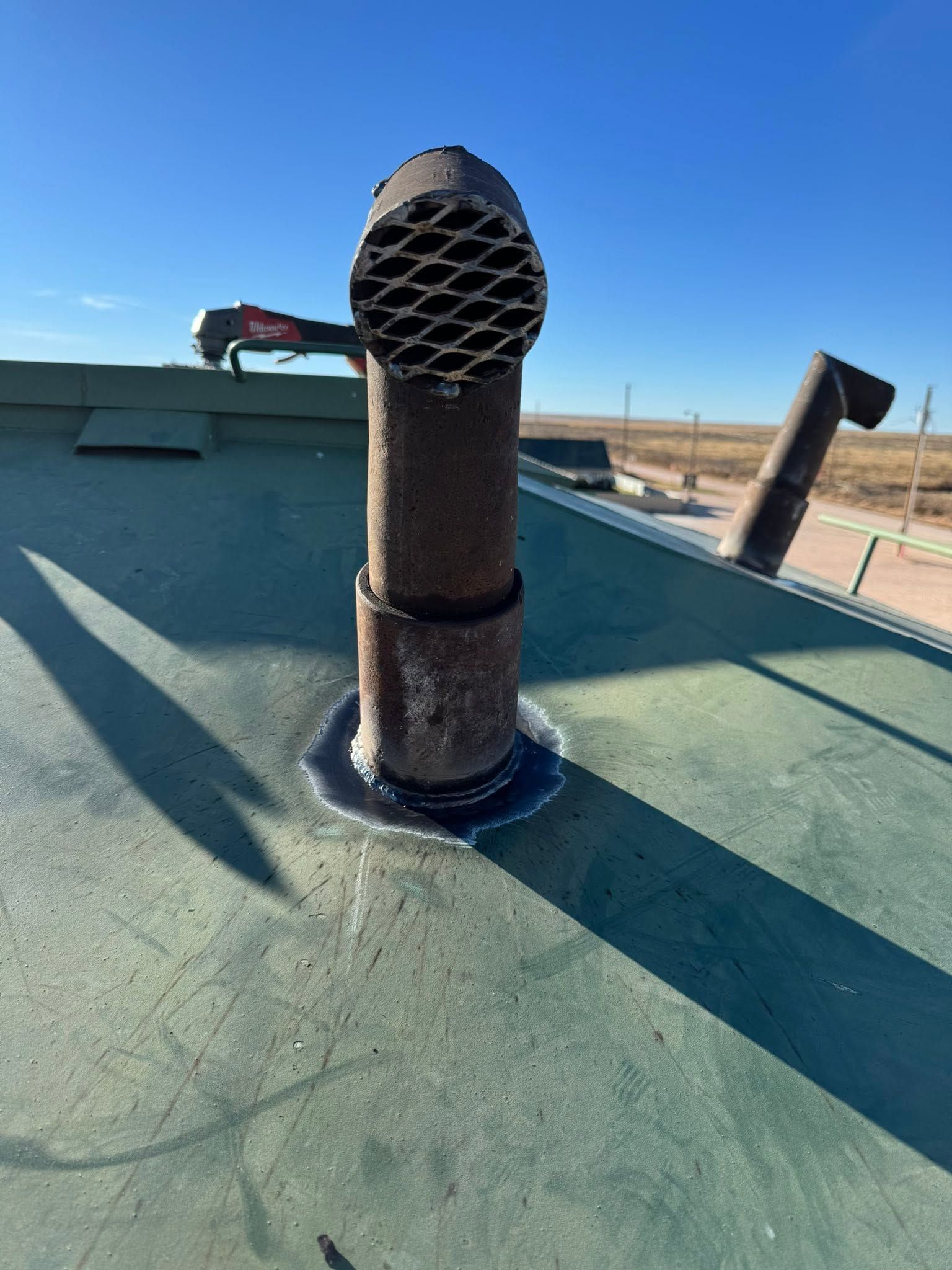 Rooftop vent pipe with black cap on green roof under a clear blue sky.