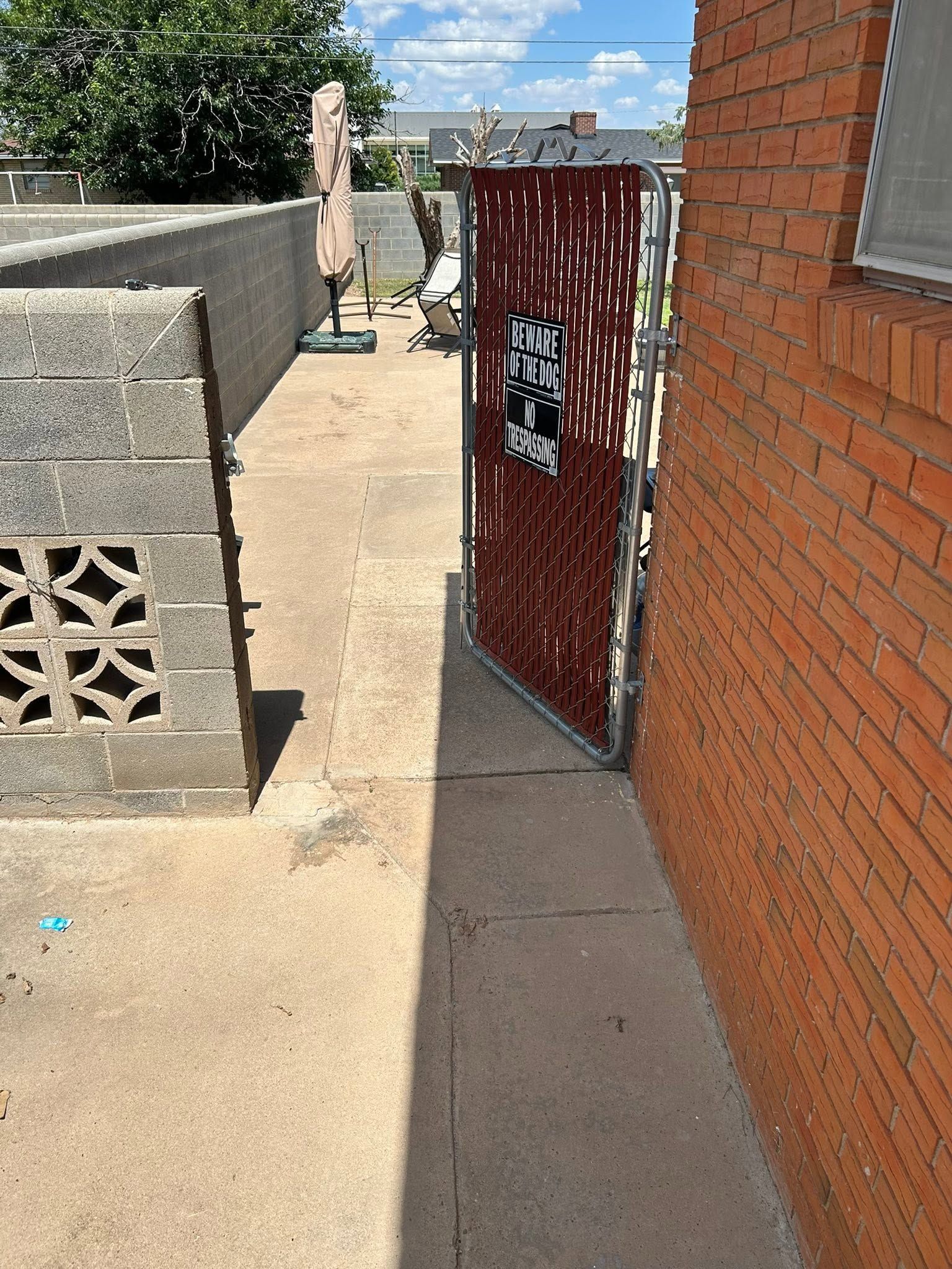 Narrow outdoor walkway beside a brick building, with a red metal gate and concrete blocks in sunlight
