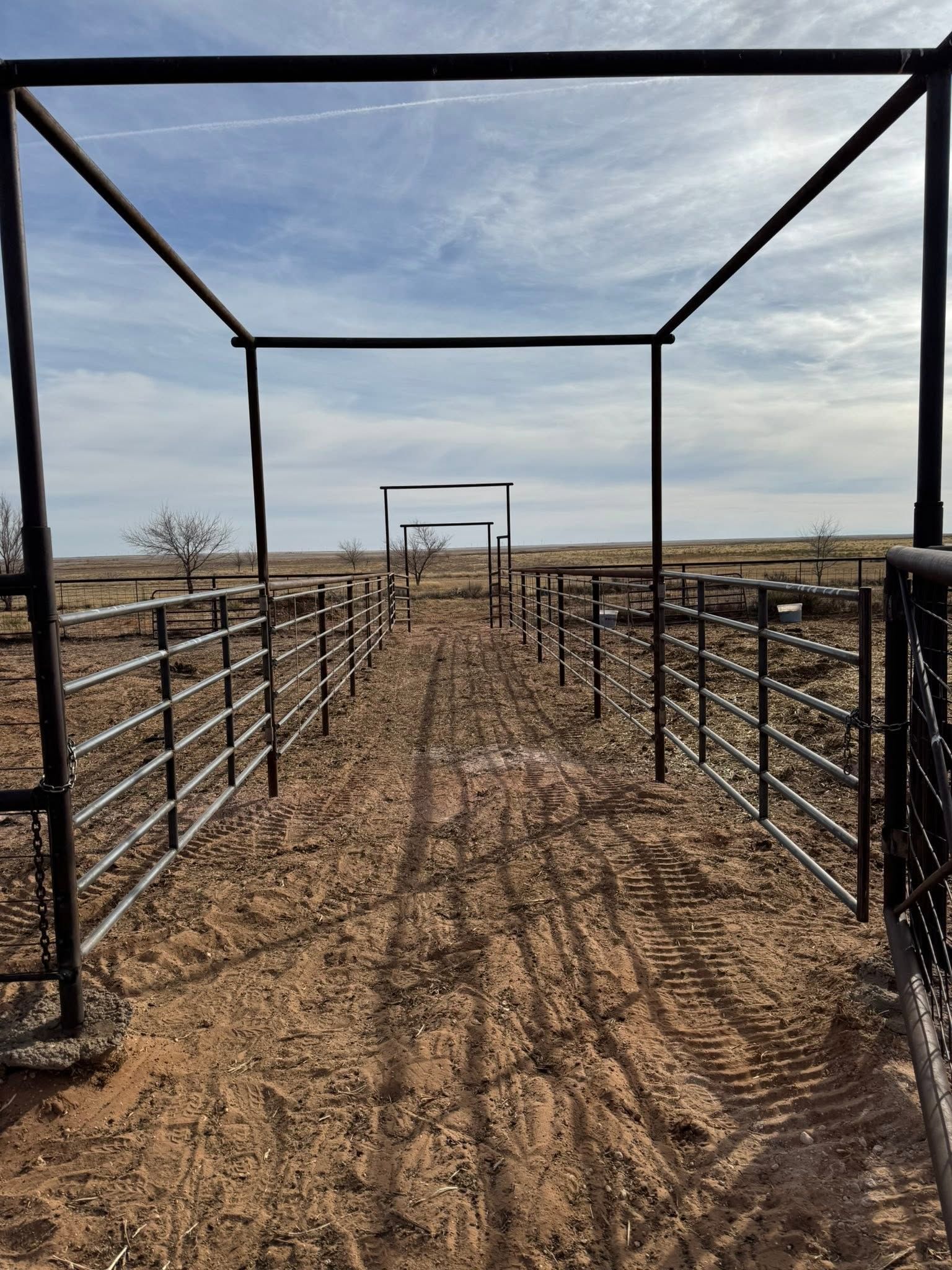 Metal livestock corral gate lined with fences in a dusty ranch landscape under a blue sky