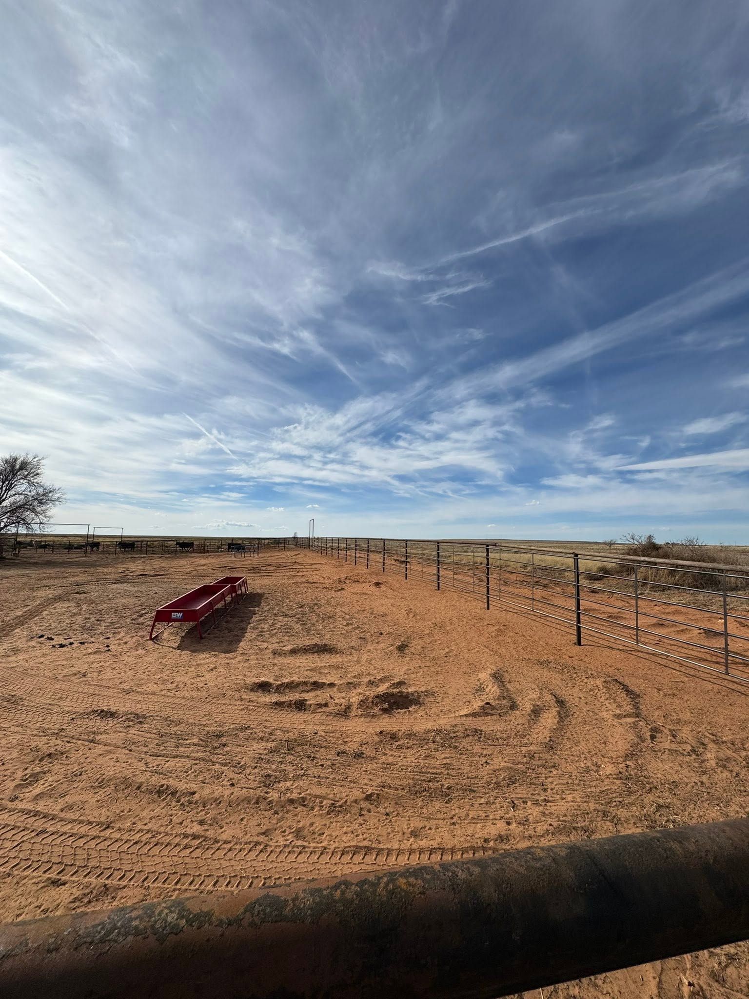 Desert roadside with a red object in the foreground, fence posts, and a wide blue sky with wispy clouds.