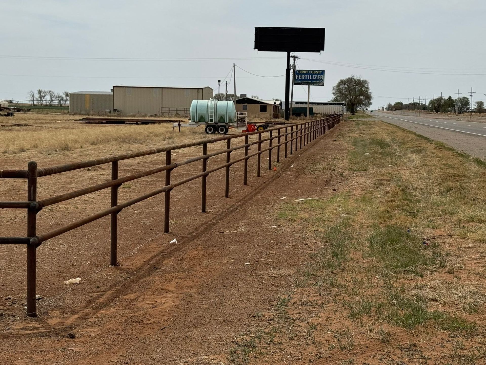 Rural roadside with rusty fence, beige buildings, utility tanks, and a highway under a cloudy sky