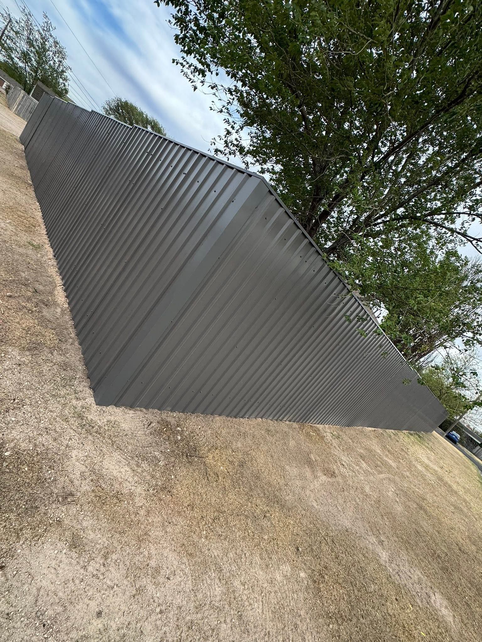Gray corrugated metal fence beside a gravel driveway, with trees overhead and blue sky visible.