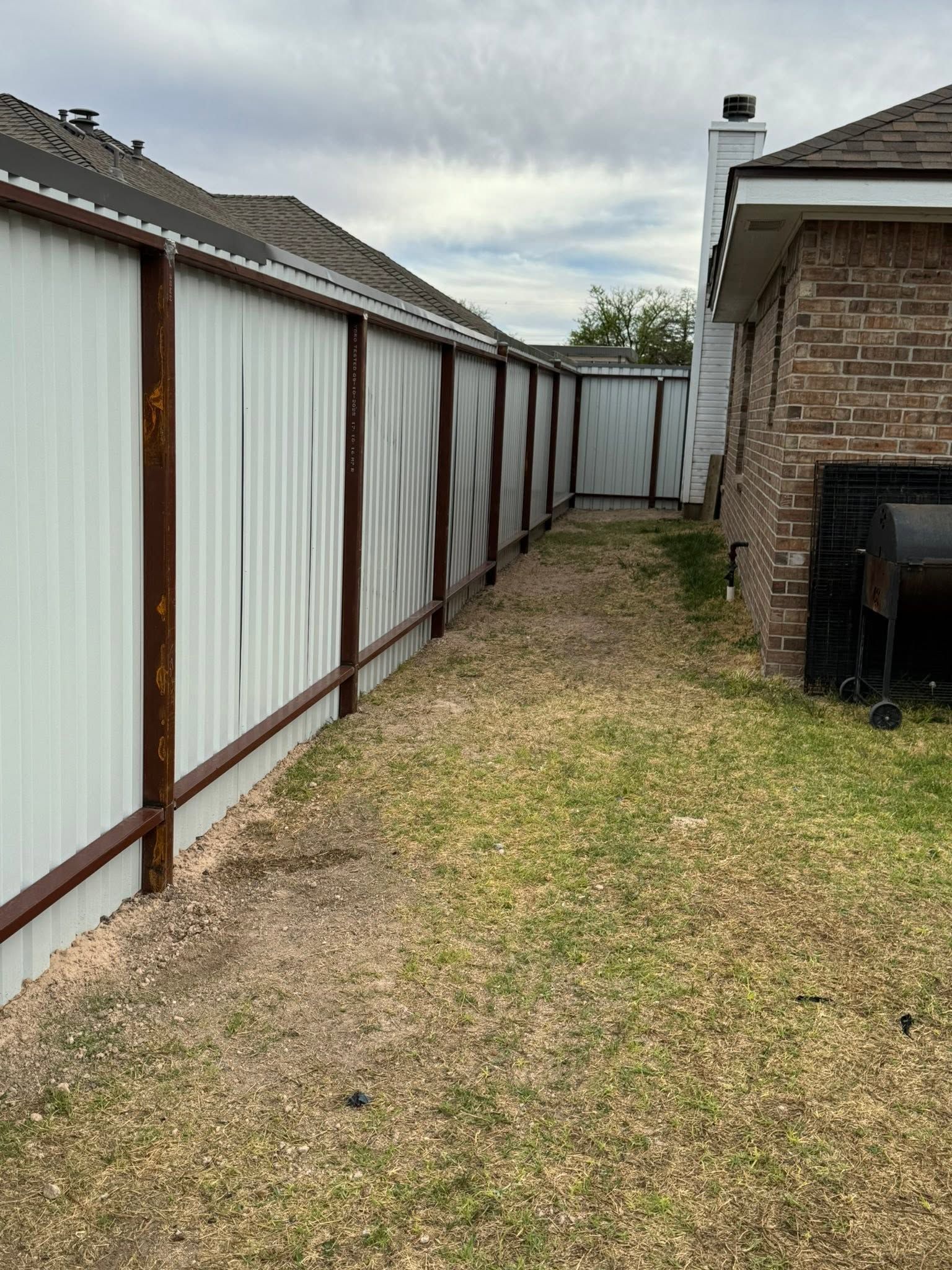 Narrow backyard between a white fence and brick house, with a small patch of grass leading to a gate.