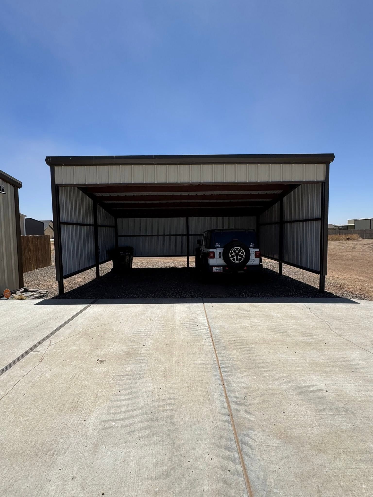 Covered metal carport with a parked vehicle, viewed from a concrete driveway under a clear blue sky